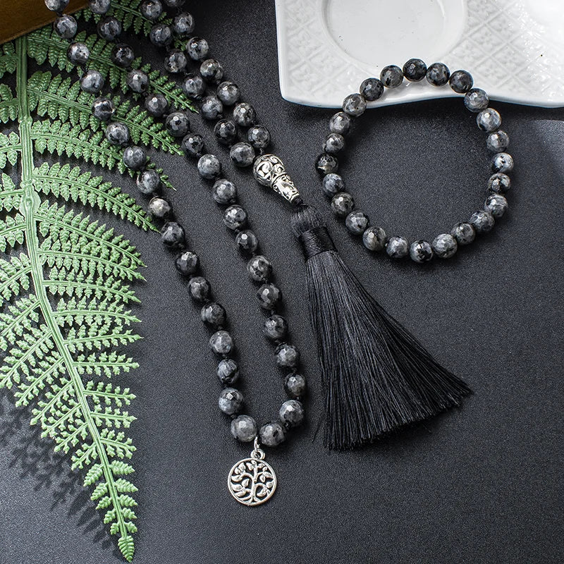 Labradorite mala, bracelet, silver charm, and black tassel arranged beside green fern leaves.